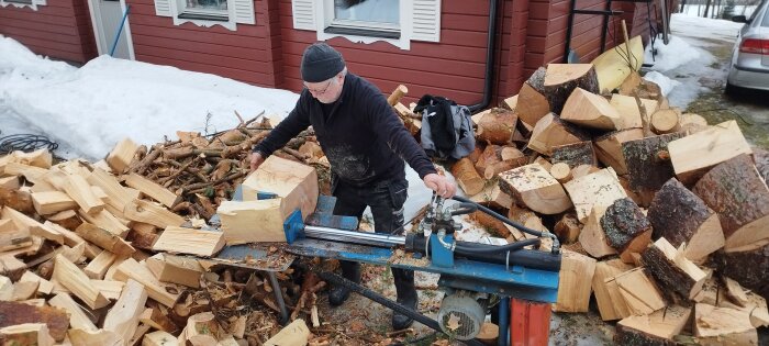 Man använder en vedklyv för att dela stockar i mindre bitar framför ett rött hus med snö runtom.