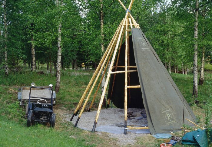 Stomme av ett kottetält byggt med trästörar och presenning i en skogsdunge, med en liten traktor bredvid.