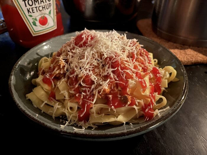 Spaghetti med köttfärssås, toppad med ketchup och riven parmesan, bredvid en flaska tomatketchup.