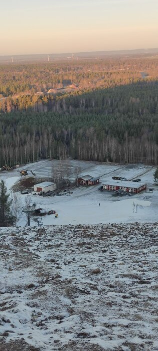 Snötäckt mark med byggnader och skog i bakgrunden, sedd från en höjd. Vindkraftverk syns i fjärran under en ljus himmel.