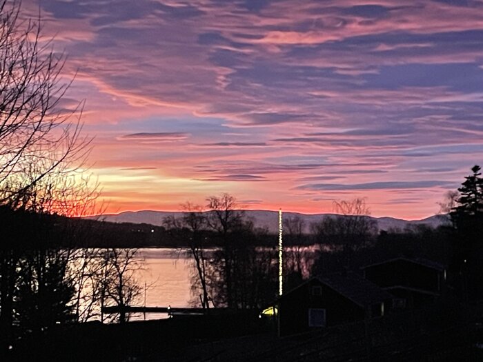 Solnedgång över en sjö med färgglada moln, siluetter av träd och byggnader i förgrunden och berg i bakgrunden.