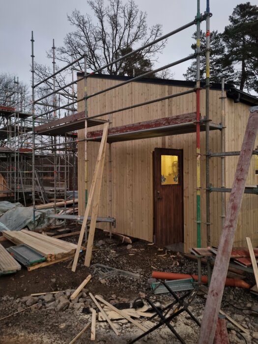 Bygge av garage med träpanel och dörr, omgiven av byggställningar och material på marken, under en mulen himmel med träd i bakgrunden.