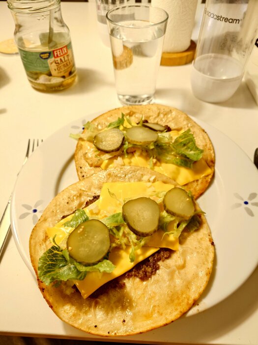 Två tortillas med ost, hamburgare, sallad och gurkskivor på en tallrik, burk med saltgurka och glas med vatten i bakgrunden.