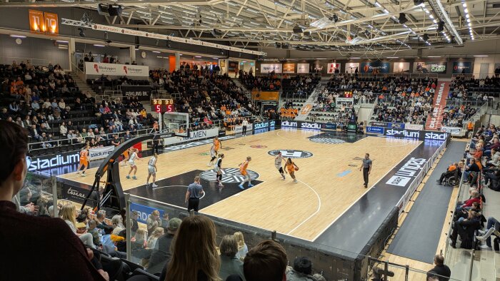 Basketmatch i en fullsatt arena, spelare i orange och vita dräkter. Publik sitter runt planen och följer spelet noggrant.