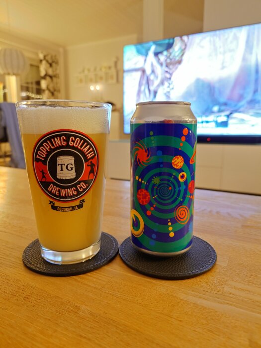 Ett glas öl från Toppling Goliath Brewing Co. och en färgglad burk öl på ett bord med en TV i bakgrunden.