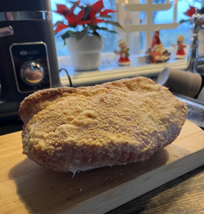 Julskinka med griljering på en skärbräda, i bakgrunden en juldekoration och en blomma i fönstret.