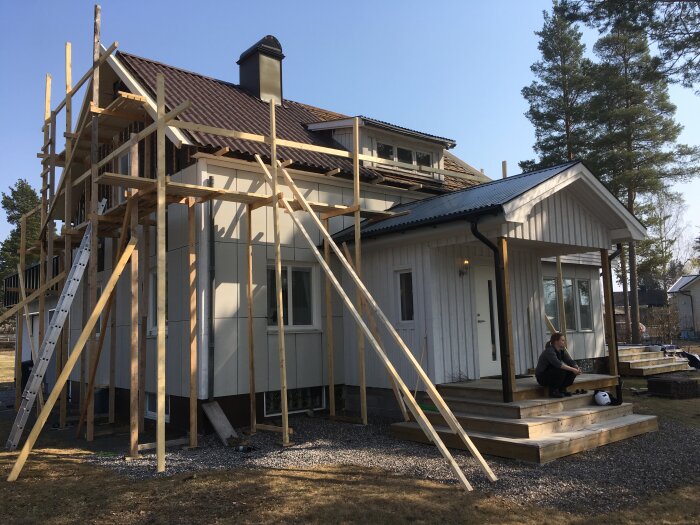 Hus från 1946 med pågående renovering av tak med byggställning, vitt träfasad och en person sitter på trappan.