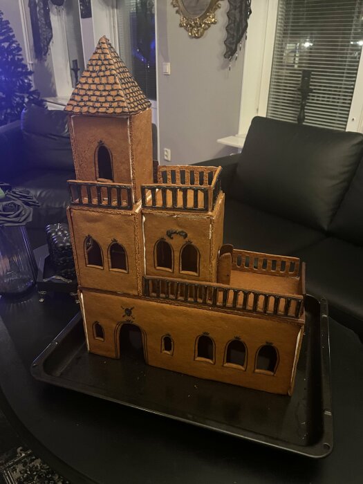 Gingerbread house shaped like a castle with a tower and detailed windows on a black tray, set in a living room.