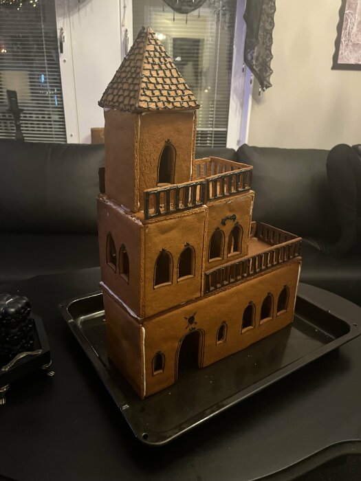 Ett pepparkakshus med flera våningar och detaljerade fönster, stående på en svart bricka i ett vardagsrum.