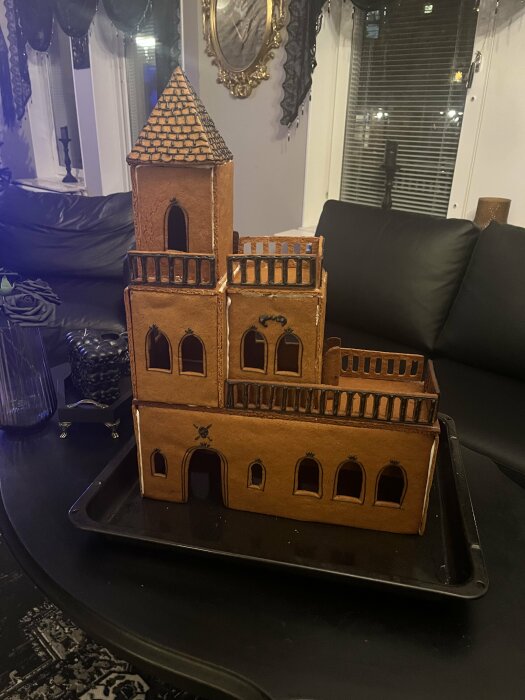 Pepparkakshus i flera våningar med torn och balkong på en svart bricka, placerat i ett vardagsrum med mörka interiörer.
