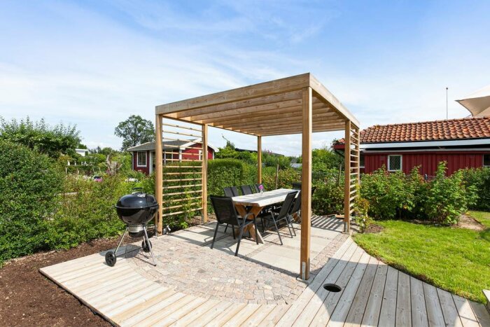 Pergola med trästomme över uteplats. Under pergolan står ett matbord med stolar, och en klotgrill syns bredvid. Grönska och rött hus i bakgrunden.