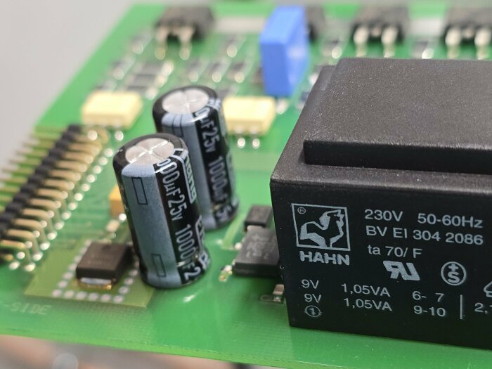 Närbild av kretskort med två elektrolytiska kondensatorer märkta "1000uF 25V" och en svart transformator märkt "HAHN 230V 50-60Hz".