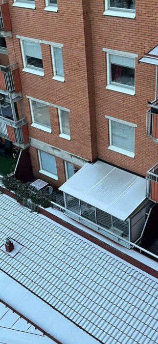 Bild av en terrass i ett flerbostadshus, delvis täckt med inglasning, omgiven av snötäckta tak och fasad i tegel.