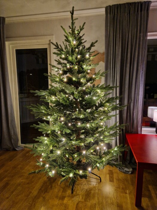 Plastgran i rum med ljusslinga, placerad framför fönster och bredvid rött bord.