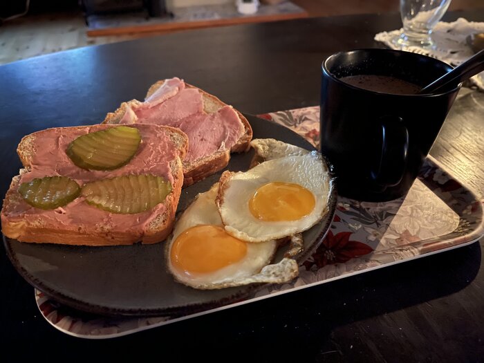 Frukost med två mackor, en med skinka och en med leverpastej och gurka, två stekta ägg och en kopp varm choklad på ett blommigt underlägg.