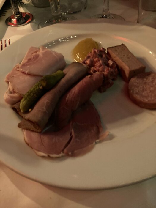 Tallrik med olika kallskuret kött, en gurka, paté och en klick senap på en vit dukad bord.