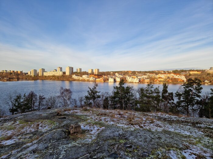 Utsikt över en sjö med flervåningshus och villor i bakgrunden, omgiven av vintergrön skog och klippor i förgrunden under en blå himmel.