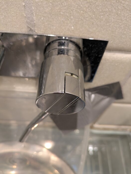 Kromad vattenkran monterad över ett handfat, med reflektioner på ytan och kakelvägg i bakgrunden.