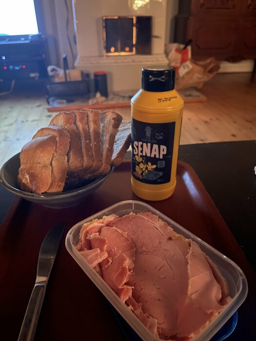 Skivat bröd, julskinka och sötstark senap på ett bord framför en brinnande kakelugn.