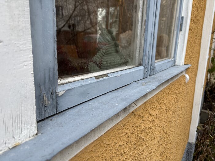 Gammalt fönster med sliten blåmålad träkarm och träfoder bredvid gul putsfasad, med kondens på glaspanelen och flagnande färg.