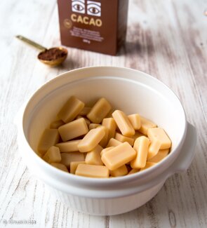Skål med ljusbruna rektangulära godisbitar på ett bord, med en låda märkt "Cacao" och sked med kakaopulver i bakgrunden.