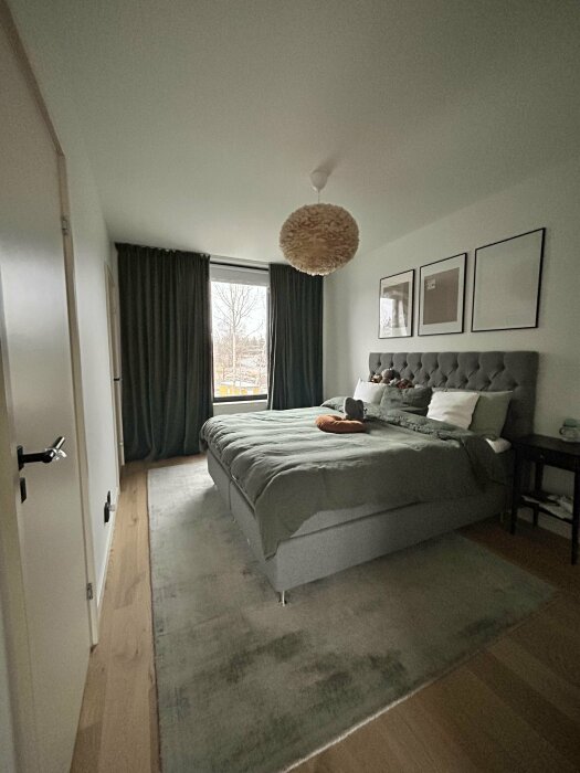 Modernt sovrum med stor grön säng, tjock matta och gardiner från golv till tak. Fönster mot utsidan, tre tavlor på väggen.