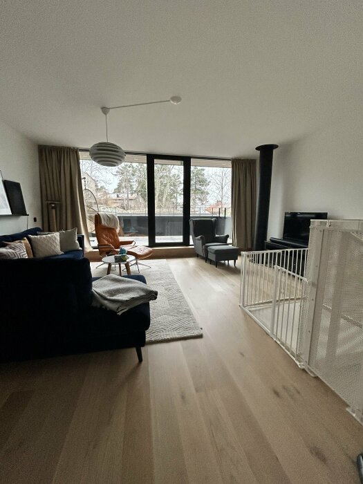 Vardagsrum med stora fönster och glasvägg mot utsidan, inredd med soffor, fåtöljer, matta och barnsäker grind, problem med buller från väg.