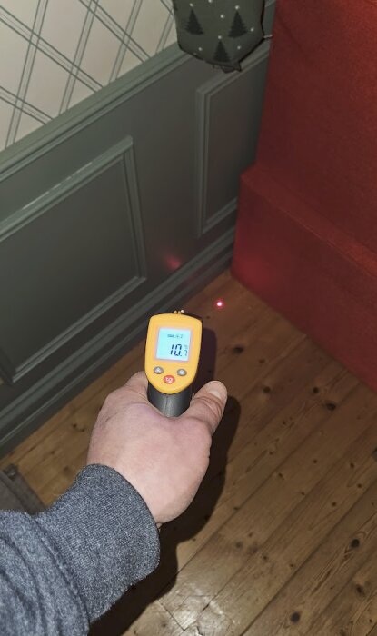 En hand håller en infraröd termometer riktad mot ett trägolv vid en vägg. Termometern visar 10,7 grader Celsius.