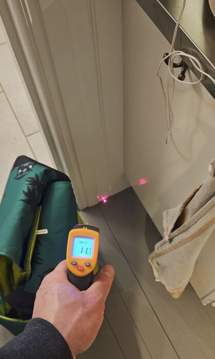 Hand håller en digital termometer som mäter 11 grader vid en vit vägg i ett rum, med laserpunkter från termometern synliga på väggen.