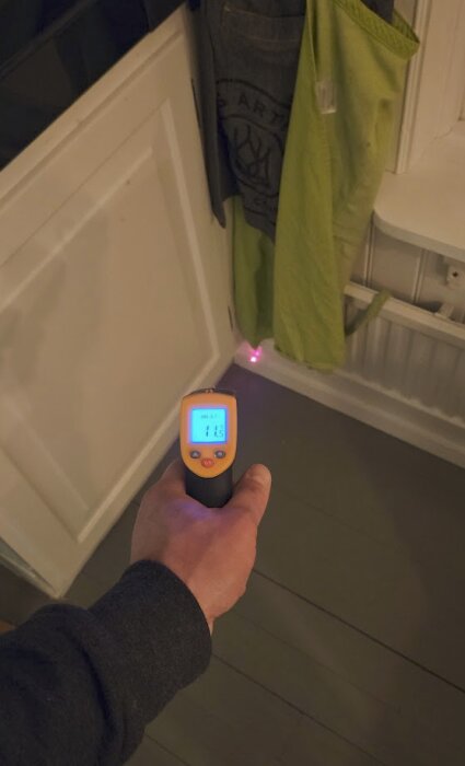 Hand som håller en IR-termometer som visar 11,5°C, med rött laserljus riktat mot ett hörn i ett rum med grått trägolv och vit panel.