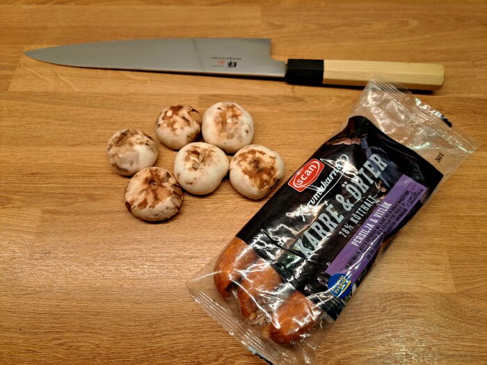 Champinjoner med bruna fläckar, en förpackning korv och en kniv på en träbänk.