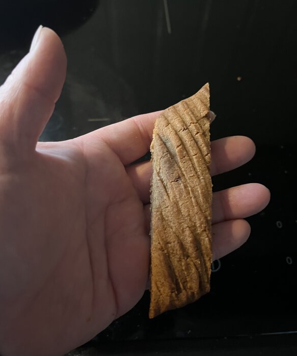 En hand håller i en kola- och pepparkakssnitt från ett recept i tidningen Hembakat. Kakan har en brun färg och är avlång med krackelerad yta.