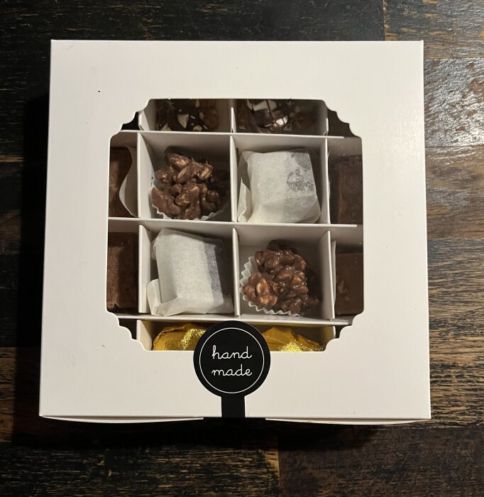 Presentask med olika hemmagjorda praliner och godbitar, i en vit låda med texten "hand made" på locket, mot en mörk bakgrund.