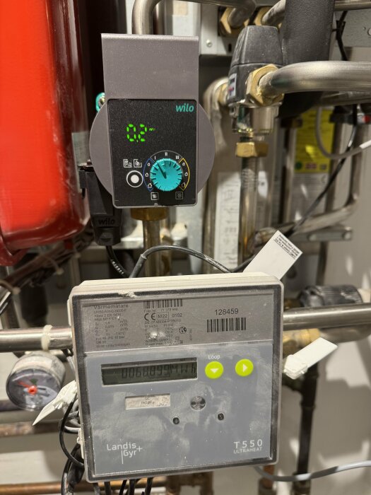 Fjärrvärmecentral med Wilo pump och Landis+Gyr T550 värmemätare, omgivna av rörledningar och en temperaturmätare.