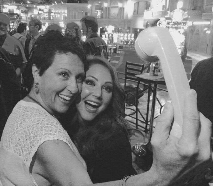 Två kvinnor poserar glatt för en bild på en livlig plats, där en av dem håller en telefonlur som en selfie-stav.