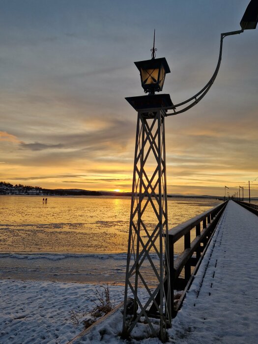 Snöig brygga vid sjön i solnedgång, med en dekorativ lampa i förgrunden och två personer långt borta på isen.