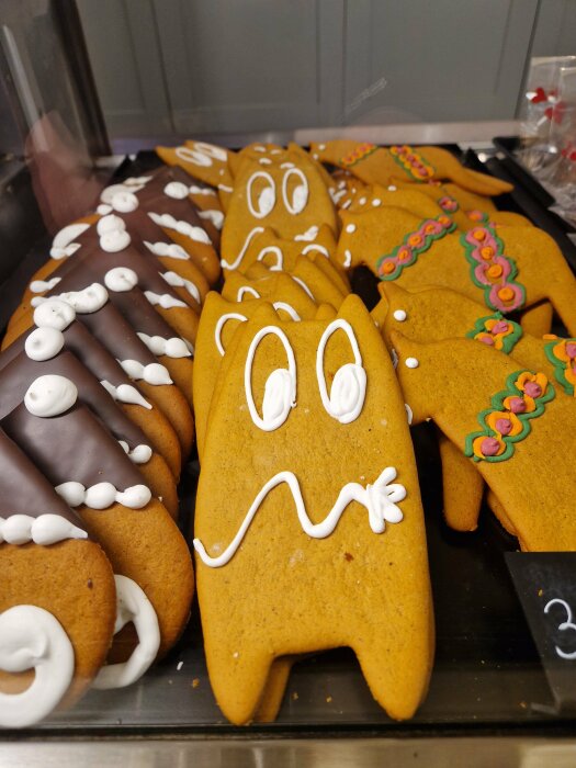 Dekorerade pepparkakor i form av Muminfigurer, med färgglada glasyrdetaljer och chokladöverdrag, på ett bageribord.