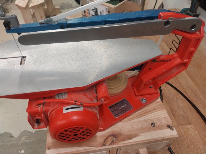 Röd och silver bandsåg monterad på en träram; Hegner Multicut-2 med motor och sågblad synligt i en verkstadsmiljö.