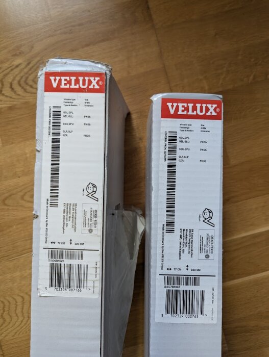 Två förpackningar märkta med Velux, innehållande eldrivna vit mörkläggningsgardin för takfönster, liggande på ett trägolv.