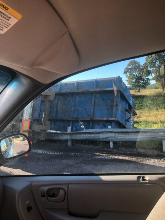 Utsikt från bilfönster mot en blå containerbil på väg bakom ett vägräcke, med gröna fält och träd i bakgrunden.