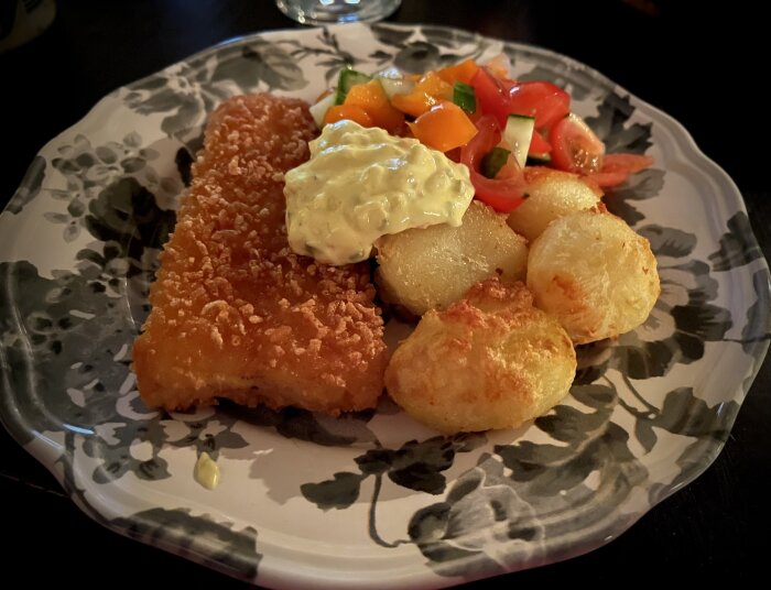 Panerad torsk med remouladsås, ugnstekt potatis och sallad på en blommig tallrik.