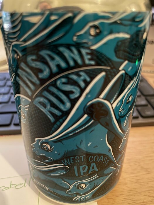 Blå burk med etiketten "Insane Rush West Coast IPA" och illustrationer av kaniner i rörelse på ett skrivbord.