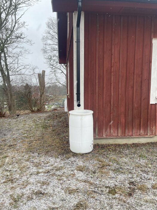 Rött hus med stuprör och vit vattentunna på sidan, snö på marken och träd i bakgrunden.