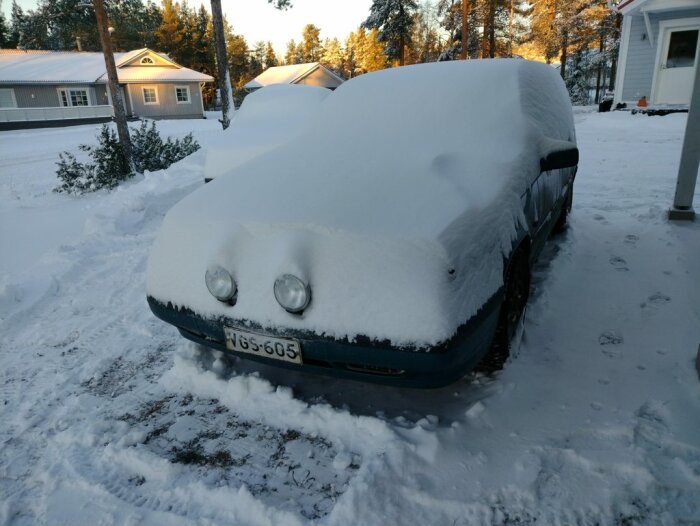 Bil täckt av snö vid vinterlandskap med hus i bakgrunden.