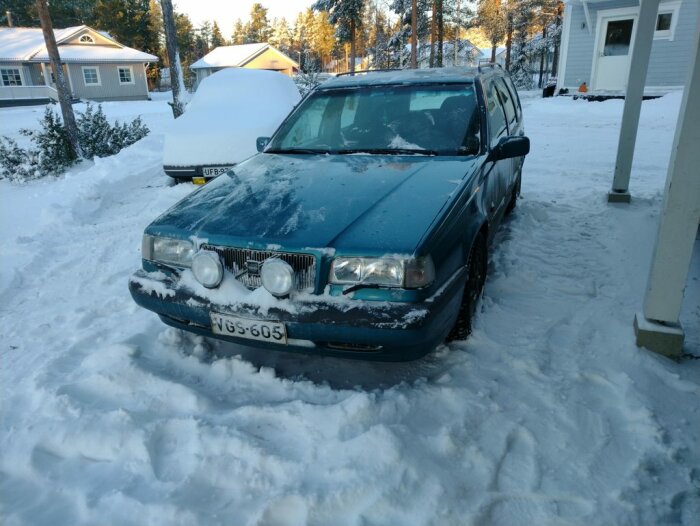 Blå bil täckt av snö parkerad på ett snöigt uppfart mellan hus, med skog i bakgrunden. Temperaturen är -14 grader.