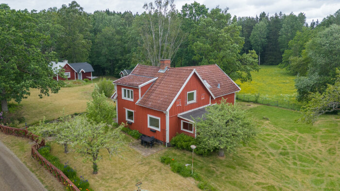 Röd träbyggnad med takkupa och tegeltak omgiven av trädgård och skog i bakgrunden. Frågor om takläggning med Plannja Trend.