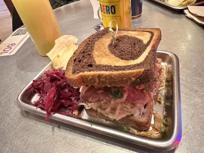 Reuben sandwich med oxbringa, surkål och rostad lök på marmorerat bröd, serverad på plåtbricka med röd lök och chips.