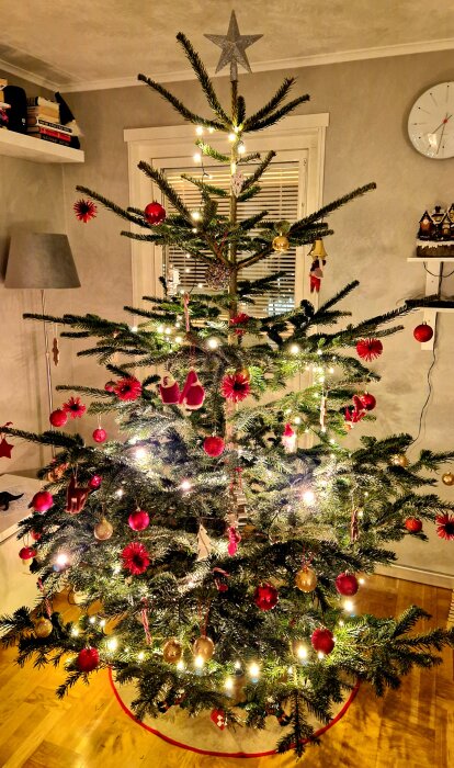 Julgran dekorerad med röda och gyllene kulor samt ljusslinga, stående på vad som beskrivs som en "Julafot", i ett hemtrevligt rum.