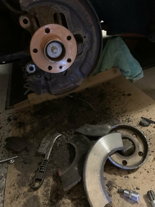 Trasig bromsskiva och verktyg på marken efter försök att byta bromsar på en bil. Smuts och rostiga delar synliga.