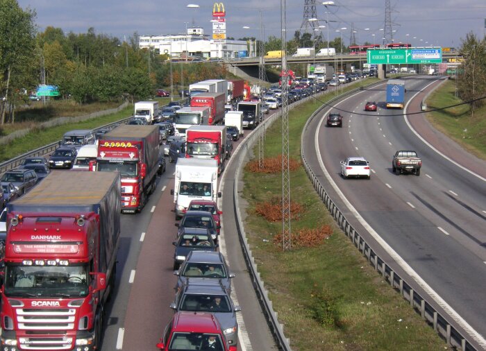 Tät trafik med lastbilar och bilar på E4 nära Stockholm, med McDonald's och andra butiker synliga i bakgrunden.